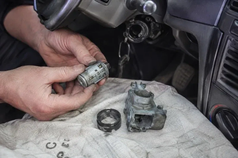 Hands holding a removed ignition cylinder beside a car steering column, with the housing and small parts on a cloth. — Ignition Lock Repair, Whitby
